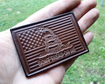 Könnte beinhalten: Ein brauner Lederaufnäher mit dem Design der amerikanischen Flagge, dem Schriftzug "DON'T TREAD ON ME" und einer zusammengerollten Schlange. Der rechteckige Aufnäher hat eine strukturierte Oberfläche und einen dunkelbraunen Rand. Ca. 7,6 cm x 5 cm.