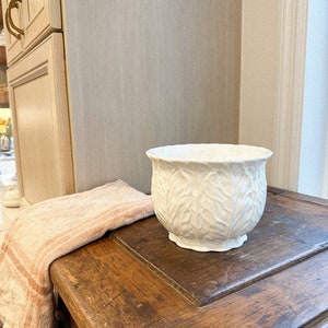 May include: A white ceramic planter with a leaf pattern sits on a weathered wooden surface. A peach and white striped towel rests nearby. The planter has a wide opening and a pedestal base. The background includes a light-colored cabinet and wall.