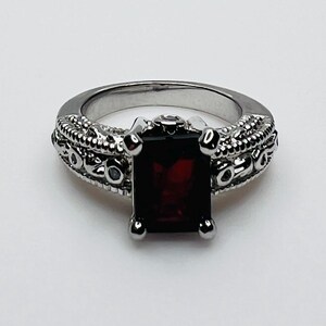May include: A silver-toned ring featuring a rectangular, deep red gemstone. The ring has an ornate, textured band with decorative detailing. The gemstone is held in place by four silver prongs.