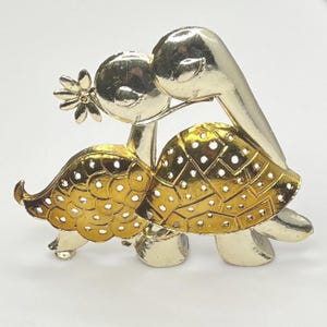 May include: A vintage gold-tone metal turtle brooch. The brooch features two turtles, one holding a flower. The turtle's shell is gold with a geometric pattern and cut-out holes. The turtles are kissing. The brooch is on a white background.