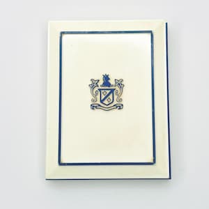 Puede incluir: Un marco de fotos rectangular blanco roto con un borde azul marino. El marco presenta un escudo azul marino en el centro. El escudo incluye una corona, un escudo y adornos decorativos. El marco es de diseño clásico.