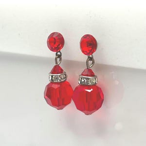 Puede incluir: Un par de pendientes de cristal rojo facetado. Cada pendiente presenta un pequeño cristal rojo redondo en la parte superior, conectado por un lazo de metal plateado a un cristal cónico rojo más pequeño. Debajo, un cristal rojo facetado más grande con una banda plateada.