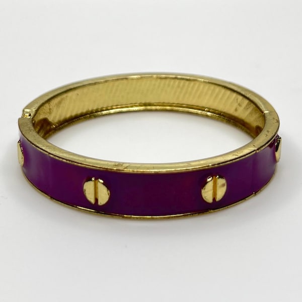 Könnte beinhalten: Ein goldfarbener Klapparmreif mit einer Einlage aus leuchtend violettem Emaille. Die Außenfläche ist mit gleichmäßig verteilten, erhabenen goldfarbenen Schraubenkopfmotiven verziert, die den gesamten Umfang des kreisförmigen Schmuckstücks umgeben.