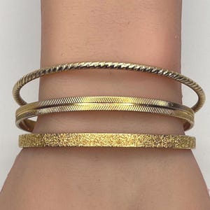 Puede incluir: Tres pulseras doradas de diferentes texturas. La pulsera superior tiene un diseño de cuerda trenzada, la del medio tiene un patrón de plumas y la inferior tiene una superficie texturizada y moteada. Estas pulseras son para la muñeca.