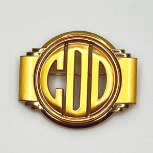 May include: A gold-coloured brooch featuring the letters "GOD" within a circular frame. The brooch has a rectangular shape with a rounded central design. It includes a pin for attachment. The design is inspired by the Art Deco style.