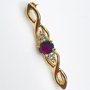 May include: A gold-toned brooch featuring an elongated, twisted metal frame. The centre is set with a single oval-shaped deep red gemstone, flanked by two clusters of three small, clear, round-cut stones positioned above and below the centre.