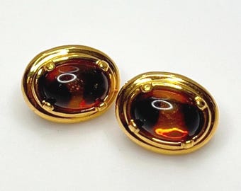 Könnte beinhalten: Ein Paar oval geformte Ohrringe mit polierten goldfarbenen Metallfassungen. Jeder Ohrring enthält einen glatten, dunkel bernsteinfarbenen Cabochon-Stein, der von vier kleinen Krappen in einem geschichteten, abgerundeten Rahmen gehalten wird.