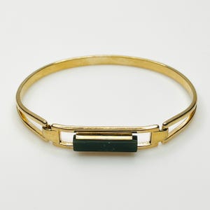 May include: A gold-toned metal bangle bracelet featuring a central rectangular dark green inlay. The bracelet has a smooth, polished finish and a hinged design with a rectangular frame that holds the green bar securely in the center.