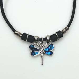 May include: A black cord necklace featuring a silver dragonfly pendant with blue and silver accents. The necklace includes black and silver beads and silver accents. The dragonfly pendant is the focal point of this jewelry piece.