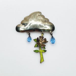May include: A silver-toned cloud-shaped brooch with two blue teardrop-shaped accents. A flower with green leaves and a brown and gold flower is suspended below the cloud. The brooch is a whimsical accessory.