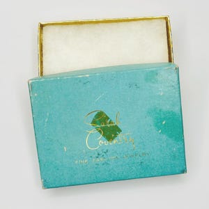May include: An open, vintage jewelry box with a teal exterior and a gold-trimmed lid. The box features the gold text "Sarah Coventry FINE FASHION JEWELRY" on the front. The interior is lined with white fabric.