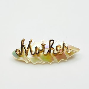 May include: A gold-coloured "Mother" hair clip. The word "Mother" is written in cursive script. The clip is set on a pearlescent, leaf-shaped base with iridescent colours of pink, yellow, and green.