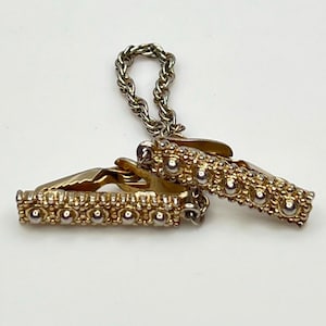 Op de afbeelding: Goudkleurige stropdasklem met een ketting en twee decoratieve staven. Elke staaf heeft een reeks kleine, zilverkleurige bollen en een getextureerd gouden oppervlak. De klem is ontworpen om een stropdas vast te zetten.