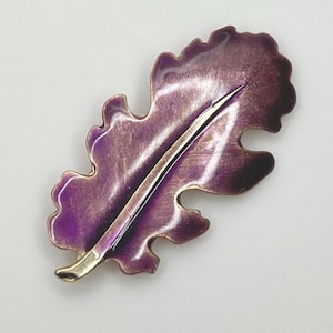 Pode incluir: Um broche em forma de folha roxa e dourada. O broche tem um design detalhado com uma veia central e uma superfície texturizada. A folha é de cor roxa escura com detalhes dourados nas bordas e no caule. O broche é provavelmente feito de metal.