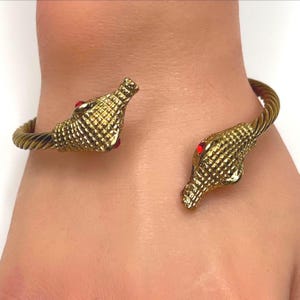 May include: Gold-toned cuff bracelet featuring snake head details with red gemstone eyes. The bracelet has a textured, twisted band and is designed to be worn open. The snake heads face opposite directions.