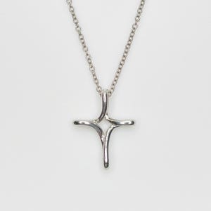 Pode incluir: Um colar com pingente prateado com um design de cruz estilizada. A cruz tem quatro braços curvos que se estendem de um ponto central, suspensos por uma corrente delicada. O colar é exibido em um fundo branco liso.