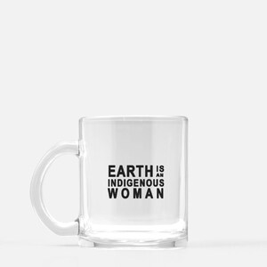 Könnte beinhalten: Transparente Glas-Kaffeetasse mit weißem Henkel. Der Becher trägt den schwarzen Text "EARTH IS AN INDIGENOUS WOMAN" in fetter, serifenloser Schrift. Geeignet für Heiß- und Kaltgetränke.