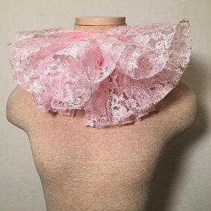 May include: A light pink lace collar with multiple ruffled layers. The collar is displayed on a mannequin and has a delicate, floral lace pattern. The collar is a soft, pastel pink color.