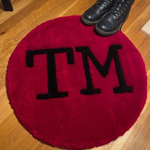 May include: A red round rug with the letters "TM" in black on it. The rug is on a wooden floor.