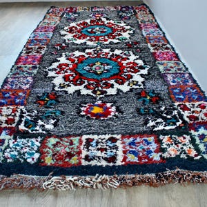 May include: A colorful Moroccan rug with a dark gray background and intricate patterns in shades of red, blue, green, and white. The rug has a fringe border and is made of wool.