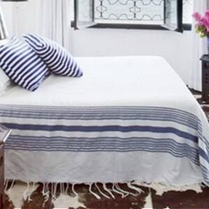 Könnte beinhalten: Weißes Bett mit einer blau-weiß gestreiften Tagesdecke und zwei blau-weiß gestreiften Kissen. Das Bett ist bezogen und die Tagesdecke ist am Fußende des Bettes gefaltet. Es gibt einen Holz-Beistelltisch mit einer Vase mit rosa Blumen darauf.