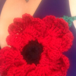 May include: A red crocheted poppy flower with a black center. The flower is on a purple background.