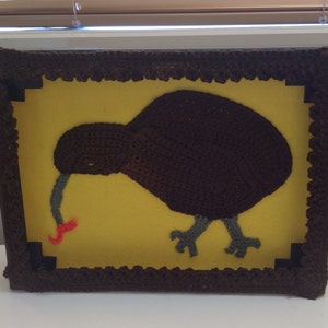 May include: A brown crocheted kiwi bird with a long beak is framed in a brown crocheted frame. The background is yellow.