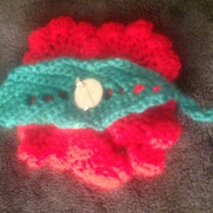 May include: A crocheted flower brooch with a red center and a teal green leaf design. A white button is attached to the leaf.