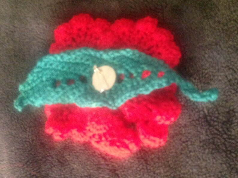 May include: A crocheted flower brooch with a red center and a teal green leaf design. A white button is attached to the leaf.