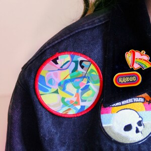 Pode incluir: Uma jaqueta de ganga preta com um remendo abstrato colorido, um remendo de caveira e dois pinos de esmalte. Um dos pinos é um coração rosa com um arco-íris e o outro diz "Angry".