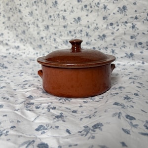 May include: A round, brown ceramic casserole dish with a matching lid and two small side handles. The pot features a smooth, glossy finish and a deep terracotta color, designed for cooking or serving food in a kitchen or dining setting.