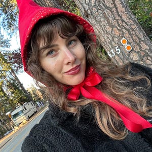 May include: A red hooded cap with white star patterns, tied with a red ribbon. The cap is worn over a black jacket. The background includes a tree trunk and a street.