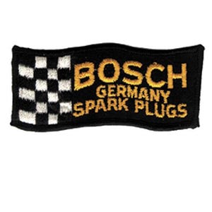 May include: A rectangular embroidered patch with a black background. The left side features a black and white checkered pattern. The right side displays the yellow embroidered text BOSCH GERMANY SPARK PLUGS in a bold, sans-serif font style.