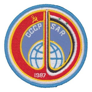 May include: A circular embroidered patch featuring a light blue centre with a white globe graphic. The text CCCP and SAR are stitched in white. A red border contains a yellow hammer and sickle, the year 1987, and a Syrian flag design element.