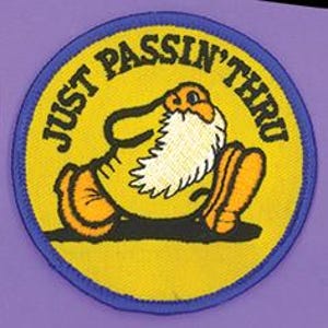 May include: A circular embroidered patch with a yellow background and a blue border. It features a cartoon character with a white beard and orange boots walking, with the black text JUST PASSIN' THRU arched along the top edge of the patch.