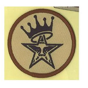 May include: A circular embroidered patch featuring a tan background with a dark brown border. The design displays a dark brown five-pointed star with a stylized face in the center, topped by a dark brown crown and a circular halo above the star.