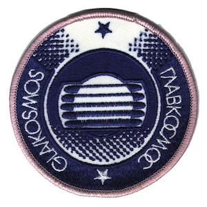 May include: A circular embroidered patch with a dark blue background and a light pink border. The center features a white sun-like graphic with horizontal lines. The text GLAVKOSMOS is written in white along the sides, with a small star at the top and bottom.