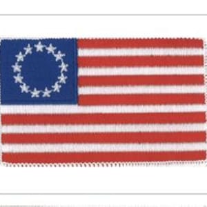 May include: A rectangular embroidered patch featuring the Betsy Ross flag design. The patch has a blue canton with thirteen white stars arranged in a circle, and thirteen alternating horizontal red and white stripes with a white stitched border.