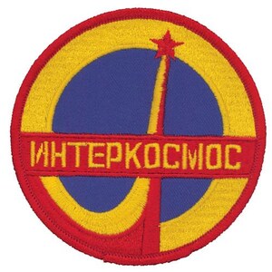 May include: A circular embroidered patch featuring a blue centre with a yellow stylised orbit and a red star at the top. A horizontal red bar across the middle displays the yellow Cyrillic text "ИНТЕРКОСМОС". The entire patch is bordered in red.