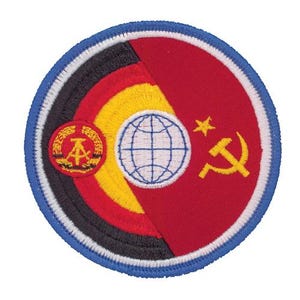 May include: A circular embroidered patch featuring a split design. The left side displays the black, red, and gold stripes of the German flag with a central emblem. The right side is red with a yellow star, hammer, and compass, plus a globe.