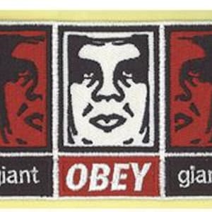 Peut inclure: Un écusson rectangulaire brodé présentant un visage blanc central sur fond noir, encadré par deux panneaux rouges avec des silhouettes noires. La partie inférieure affiche le texte giant en blanc, OBEY en blanc sur rouge et giant en blanc.