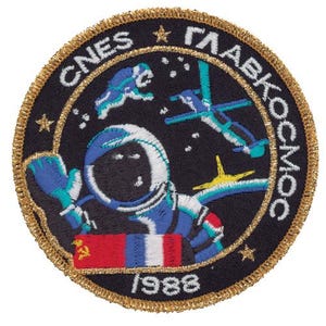 May include: A circular embroidered patch with a black background and gold border. It features an astronaut in a blue and white suit, a space station, stars, and the text CNES, GLAVKOSMOS, and 1988. A rectangular flag with Soviet and French stripes is visible.