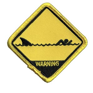 Könnte beinhalten: Ein rautenförmiger gestickter Aufnäher mit gelbem Hintergrund und schwarzem Rand. Das Design zeigt eine schwarze Grafik einer Haifischflosse und eines Schwimmers im Wasser, mit dem Wort WARNING in schwarzen Großbuchstaben am unteren Rand.