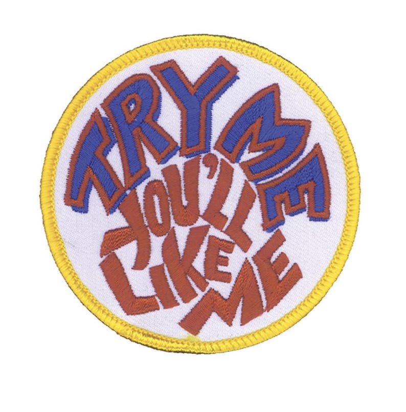 Puede incluir: Un parche bordado circular con fondo blanco y borde amarillo. El texto "TRY ME YOU'LL LIKE ME" est&aacute; cosido en el centro, con "TRY ME" en azul y "YOU'LL LIKE ME" en color rojo-marr&oacute;n, todo en una fuente negrita y estilizada.