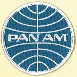 May include: A circular embroidered patch featuring a blue background with white stitched lines forming a globe grid pattern. The text PAN AM is embroidered in white capital letters across the centre of the patch, which has a white border.