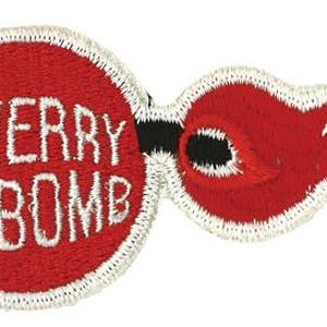 Könnte beinhalten: Ein gestickter Aufnäher, der eine kreisförmige rote Kirsche mit dem weißen Text CHERRY BOMB auf der Oberfläche zeigt. Auf der rechten Seite befindet sich ein rotes, flammenförmiges Element mit schwarzer Mitte und weißem Rand, das komplett mit weißem Garn umrandet ist.
