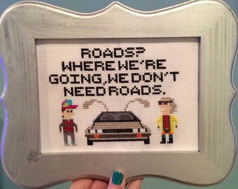 May include: A framed cross-stitch featuring a scene from the movie Back to the Future. The text reads "Roads? Where we're going, we don't need roads."