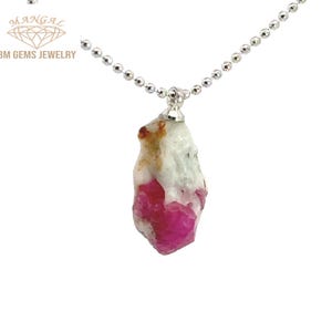 May include: A raw ruby pendant necklace. The pendant is a rough, irregular shape with a vibrant pink and white color. The necklace chain is a delicate silver ball chain. The brand name "BM GEMS JEWELRY" is visible in the upper left corner.