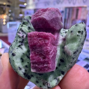 May include: A natural ruby crystal cluster, showcasing a deep red hue, is set on a green stone base with black speckles. The rough-cut ruby crystals are the focal point, highlighting the raw beauty of the gemstone.