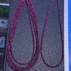 Può includere: Due collane con piccole perline rotonde rosso scuro. Una collana è a filo singolo, l'altra a più fili. Le collane sono esposte su una superficie blu scuro strutturata.